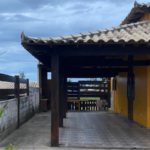Casa à venda na Praia de João Francisco, em Quissamã. Foto da frente da casa amarela, com detalhes em madeira, com quintal, garagem e jardim.
