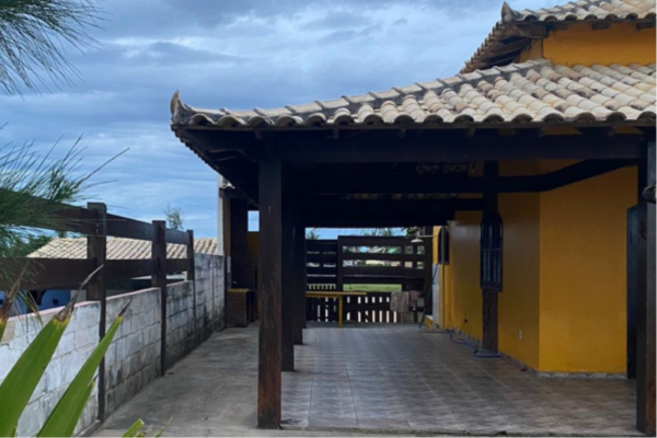 Casa à venda na Praia de João Francisco, em Quissamã. Foto da frente da casa amarela, com detalhes em madeira, com quintal, garagem e jardim.